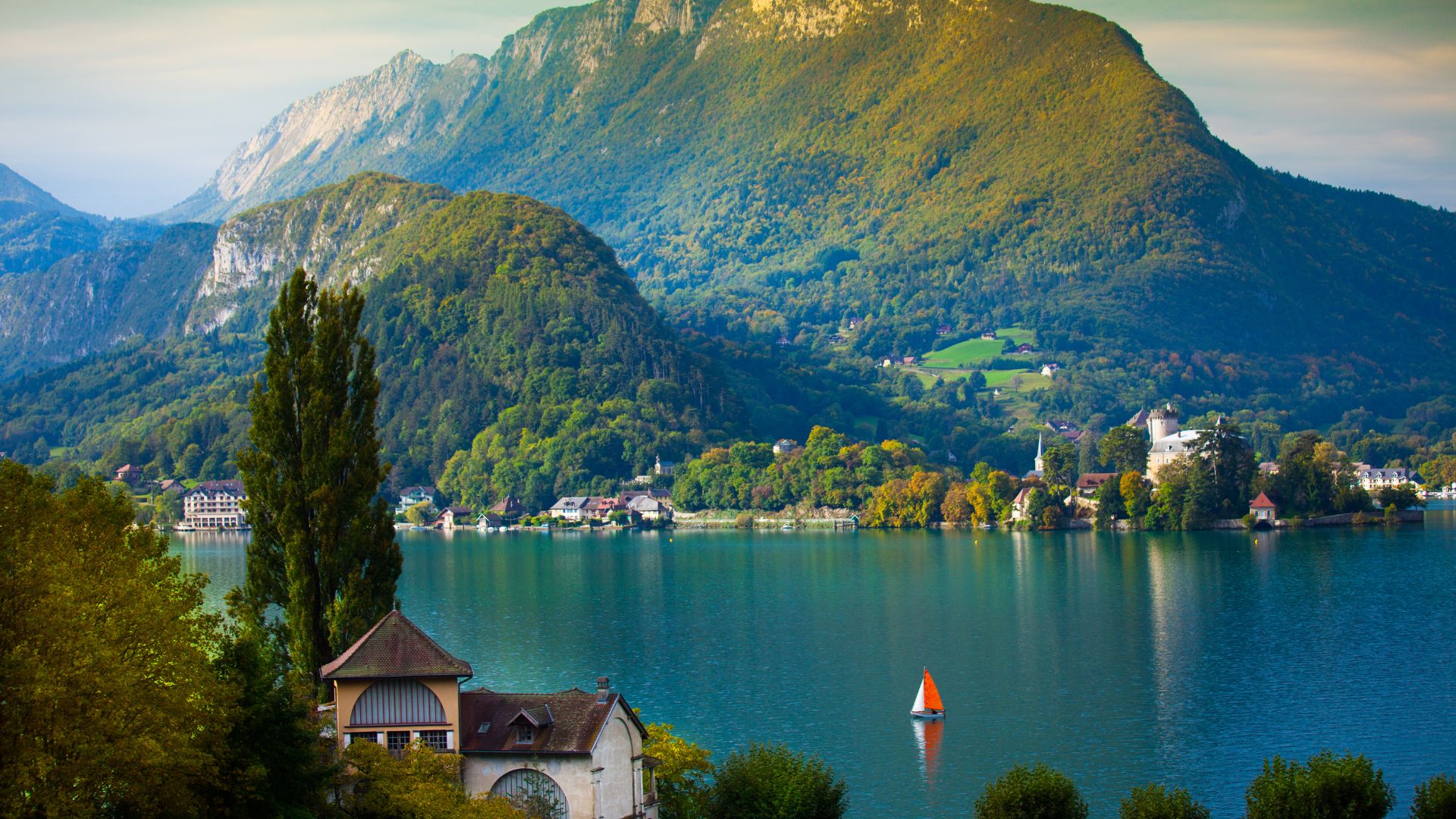 Lac d’Annecy en Haute-Savoie Vue panoramique sur le lac d’Annecy entouré de montagnes verdoyantes en Haute-Savoie, avec un voilier rouge et blanc naviguant sur des eaux calmes, et un château visible au loin, en bord de rive.